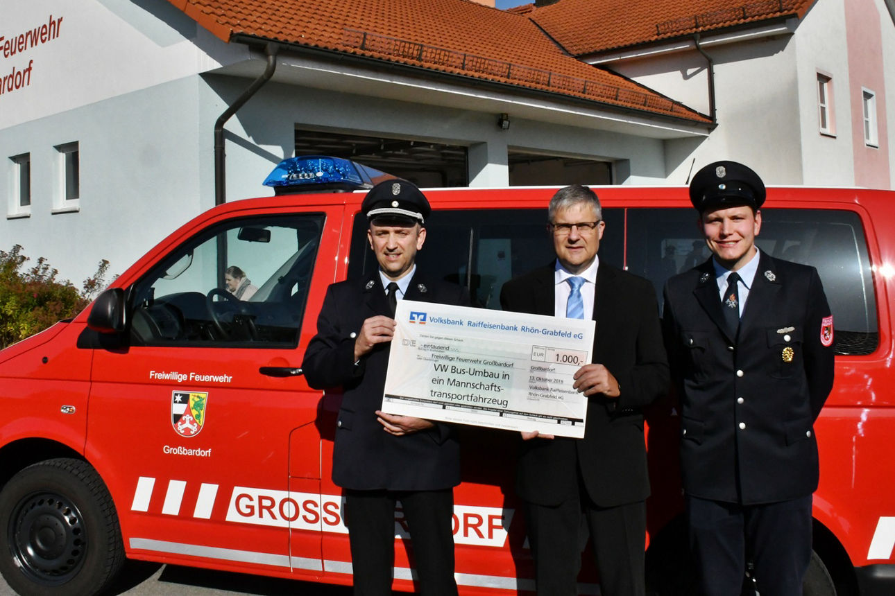 Drei Männer stehen vor einem Feuerwehrauto. Zwei Männer halten einen Spendenscheck in der Hand 
