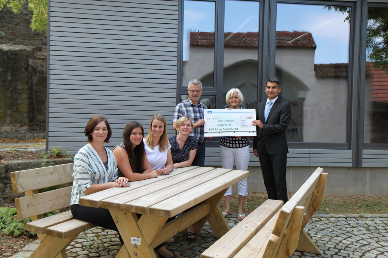 Auf einer Holzbank sitzen vier Frauen, dahinter stehen zwei Männer und eine Frau, die einen Spendenscheck halten 