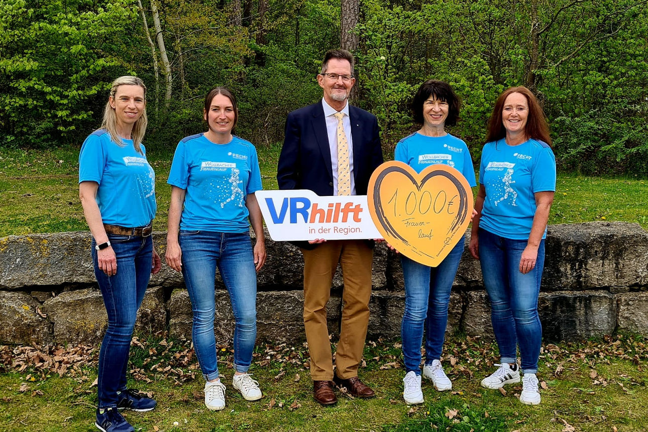 Fünf Personen stehen draußen vor einer Steinmauer sie halten ein großes orangefarbenes Herz mit der Aufschrift „1.000 € Fraue