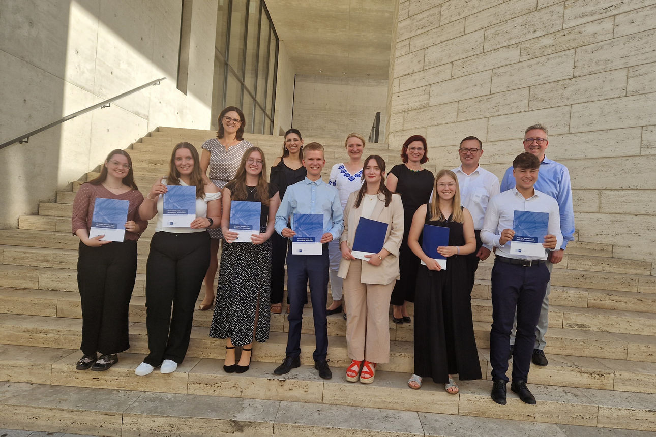 Sieben junge Menschen stehen auf einer Treppe und halten ein Zeugnis in der Hand, hinter ihnen vier Frauen und zwei Männer