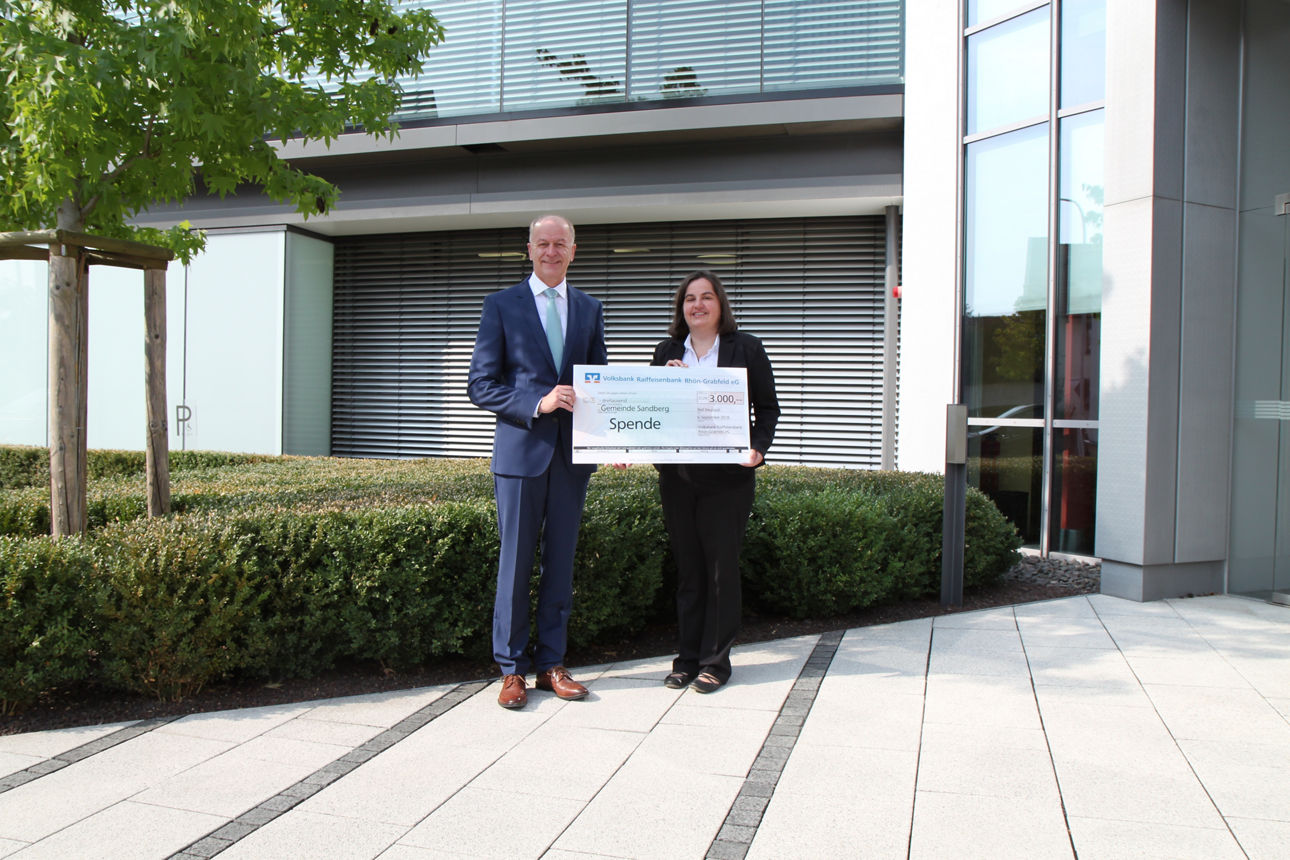 Ein Mann und eine Frau stehen vor einem Gebäude und halten einen Spendenscheck in der Hand 