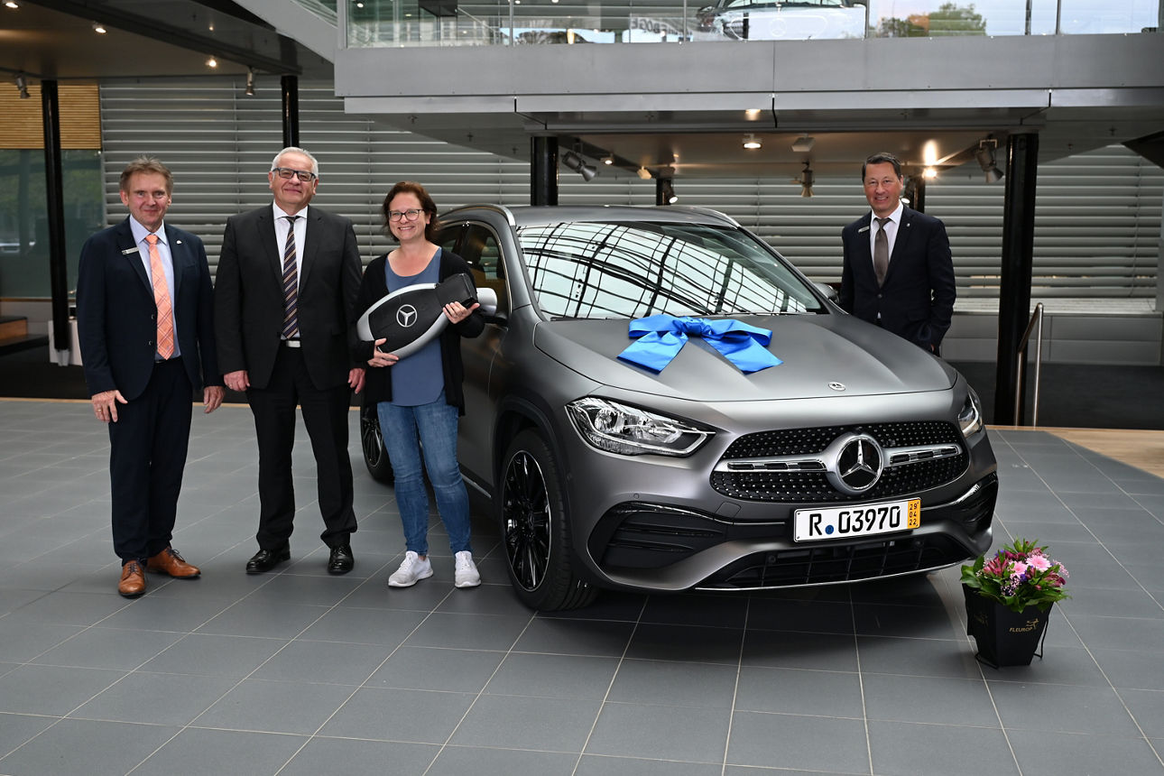 Vier Personen stehen in einer Autohalle neben einem grauen Mercedes mit einer großen blauen Schleife