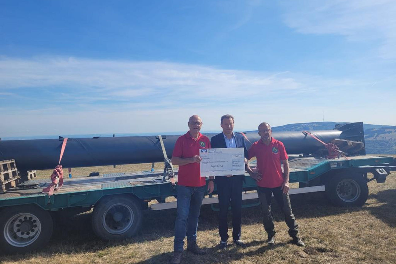 Drei Männer stehen vor einem Anhänger, zwei von Ihnen halten einen Spendenscheck 