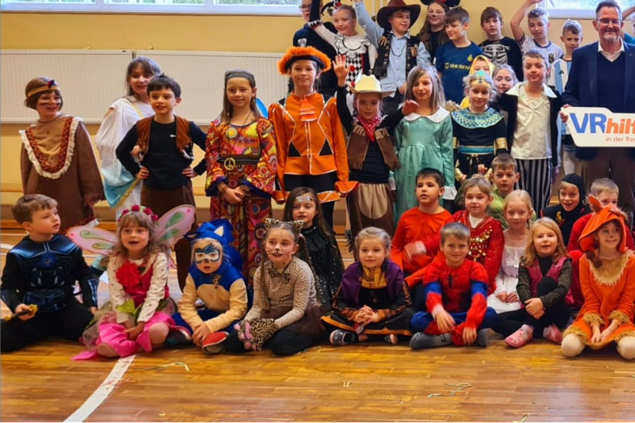 Eine große Gruppe von Kindern in bunten Kostümen sitzt und steht in einer Turnhalle, begleitet von zwei Erwachsenen