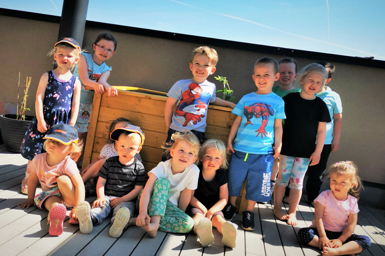 Eine Gruppe von kleinen Kindern steht und sitzt lachend auf einer Terrasse bei sonnigem Wetter, mit einem hölzernen Hochbeet 