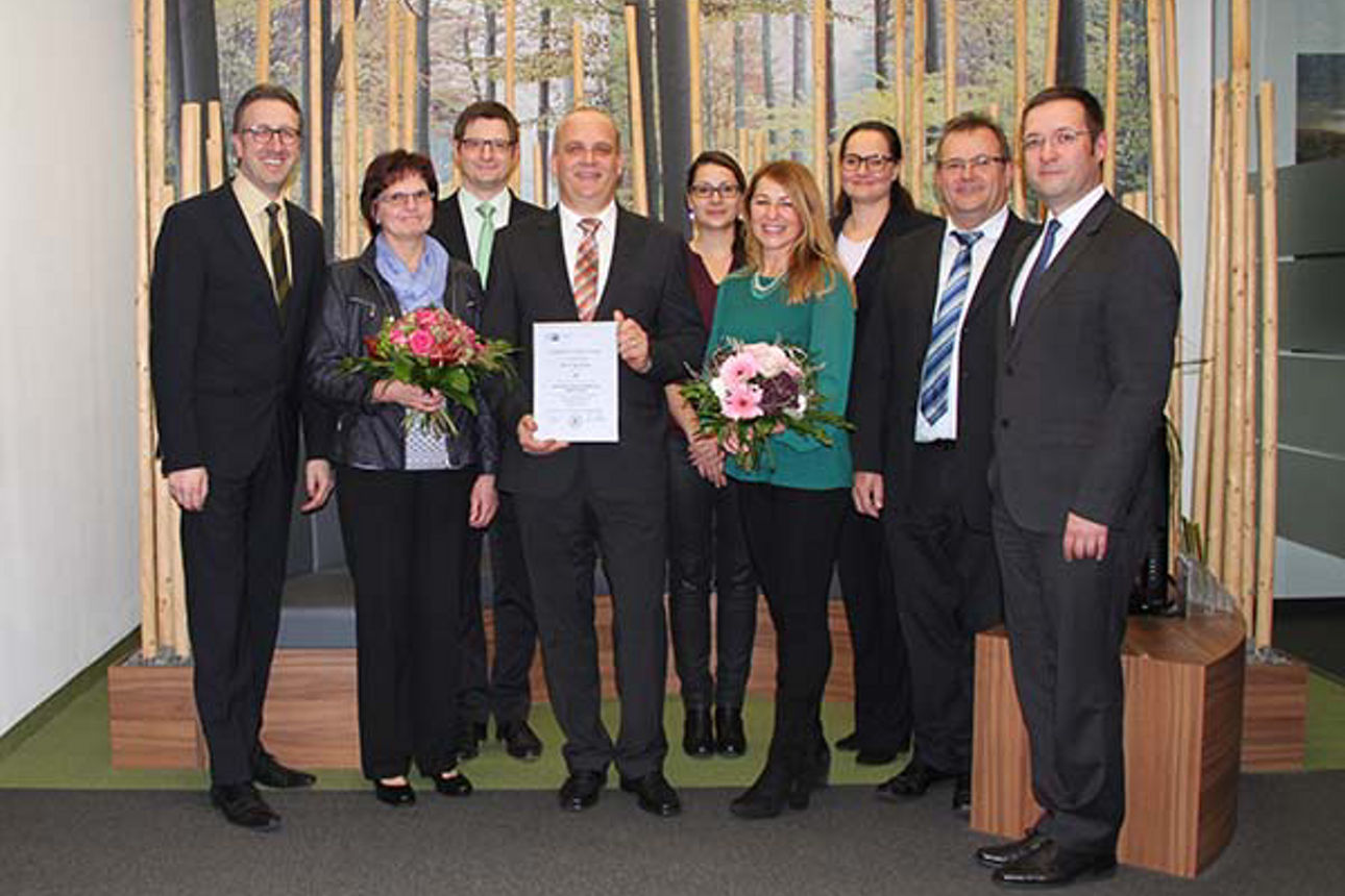 Einige Menschen stehen vor einer Wand mit Waldmotiv, zwei von ihnen halten Blumen und ein Mann eine Urkunde 