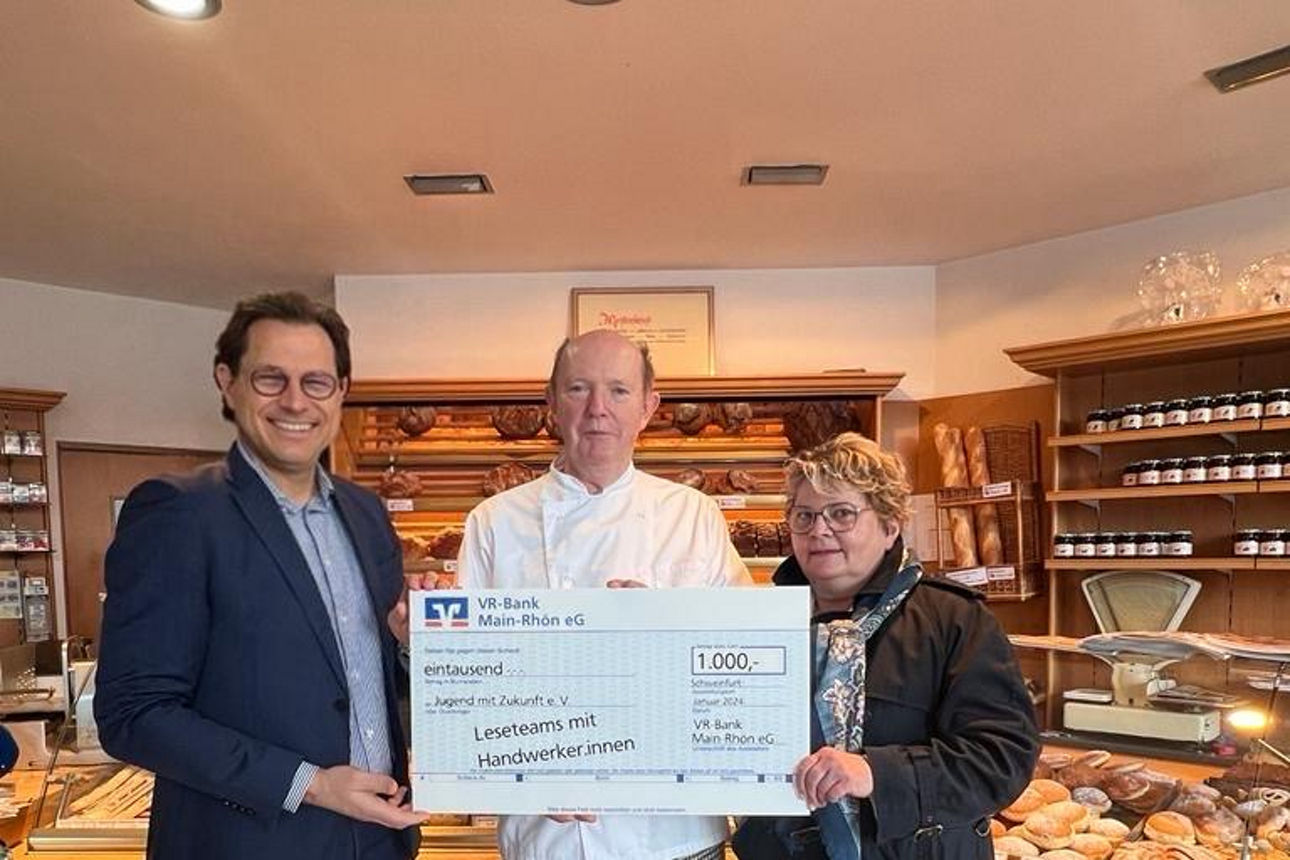 Drei Personen stehen in einer Bäckerei, halten gemeinsam einen symbolischen Scheck über 1.000 €