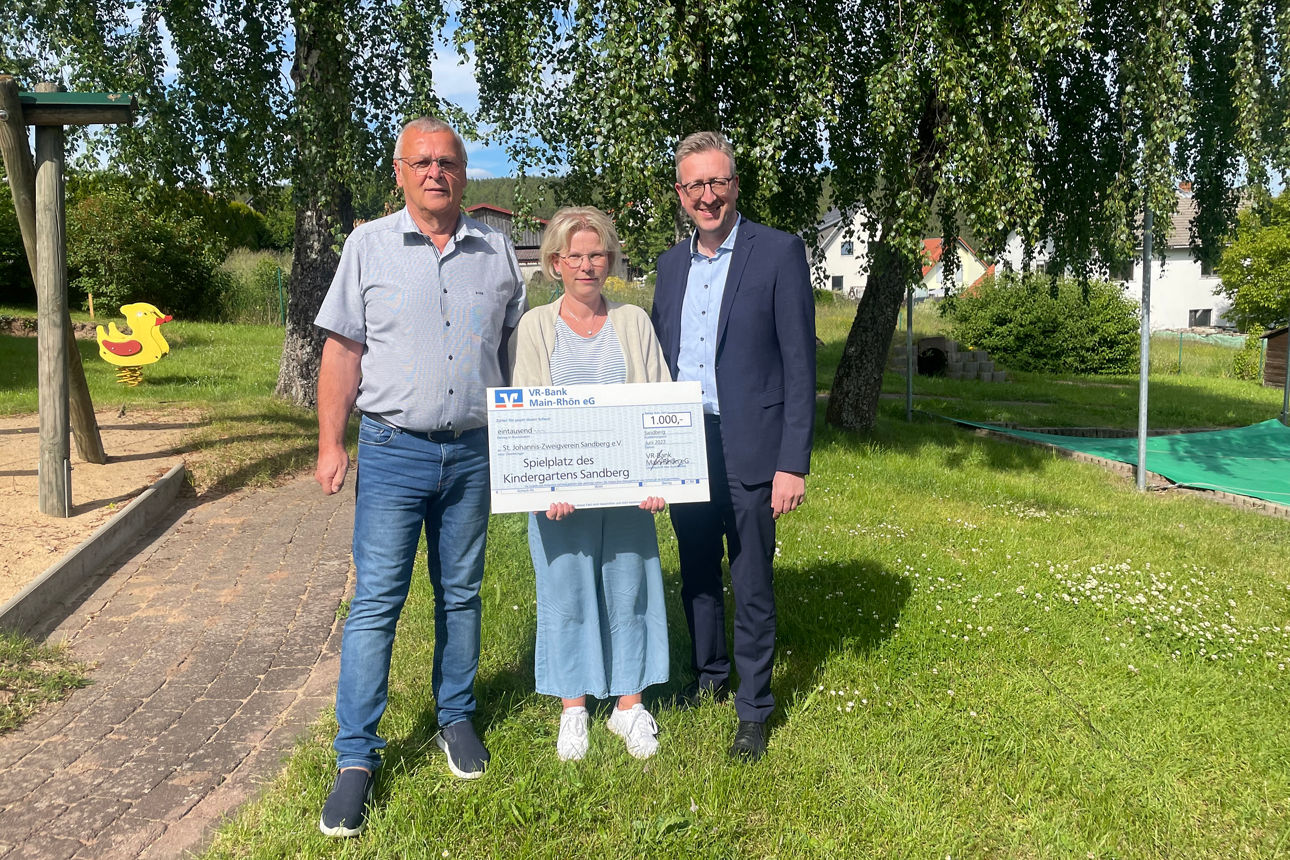 Drei Personen stehen auf einer Wiese vor einem Spielplatz und halten einen Spendenscheck über 1.000 Euro für den Kindergarten