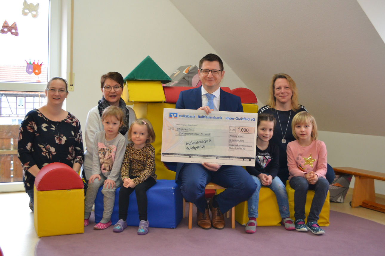 Links sitzen zwei Frauen und zwei Kinder, rechts eine Frau und zwei Kinder, ein Mann der einen Spendenscheck in der Hand häl