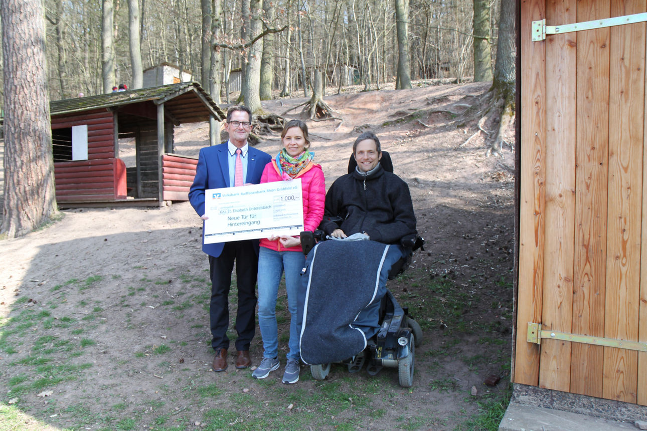 Ein Mann im Rollstuhl, ein Mann der steht und eine Frau beide einen Spendenscheck haltend stehen neben einem Holzhaus.