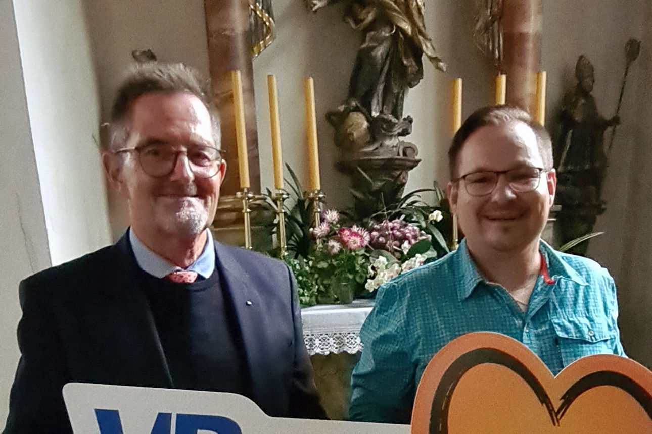 Zwei Männer stehen in einer Kirche vor einem geschmückten Altar, halten ein Schild mit der Aufschrift „VRhilft in der Region“