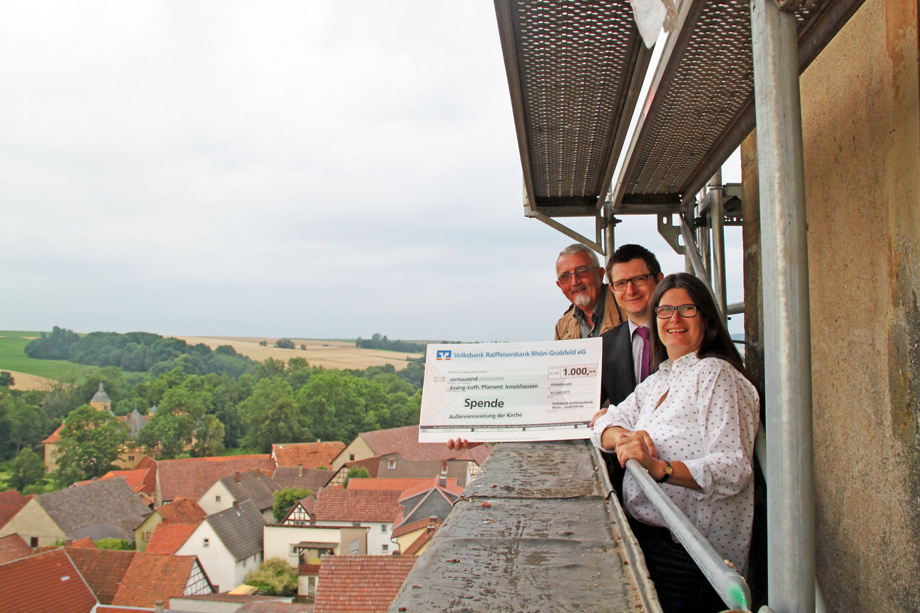 Zwei Männer und eine Frau stehen auf einer Erhöhung, wobei ein Mann einen Spendenscheck in der Hand hält.