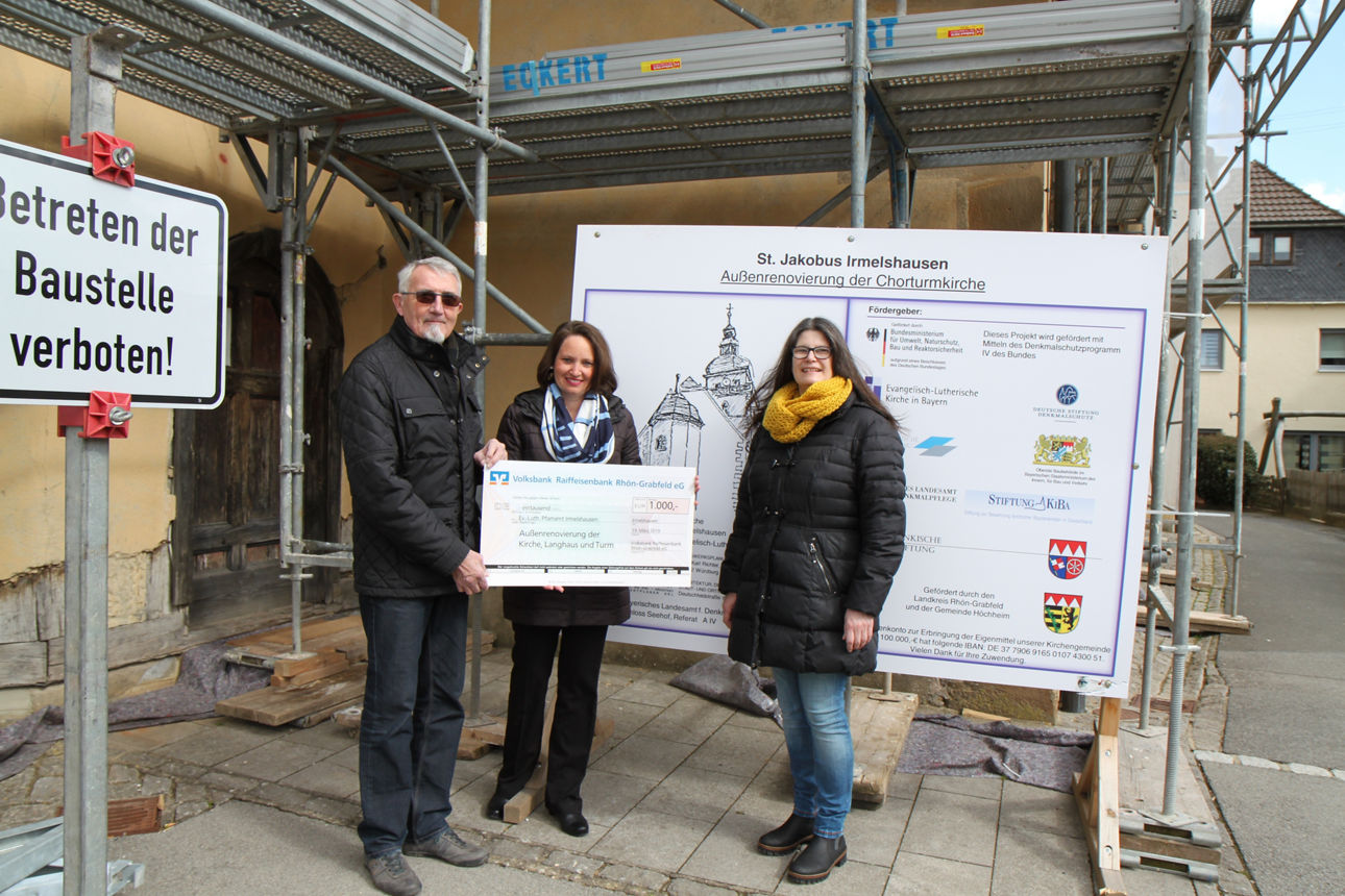 Ein Mann und zwei Frauen stehen vor einer Bautafel wobei der Mann und eine Frau einen Spendencheck in der Hand hält 