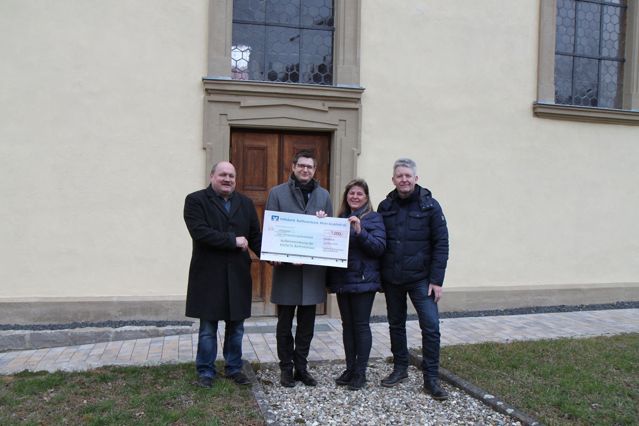 Drei Männer und eine Frau stehen vor einem Gebäude. Zwei Männer und die Frau halten einen Spendenscheck in der Hand 