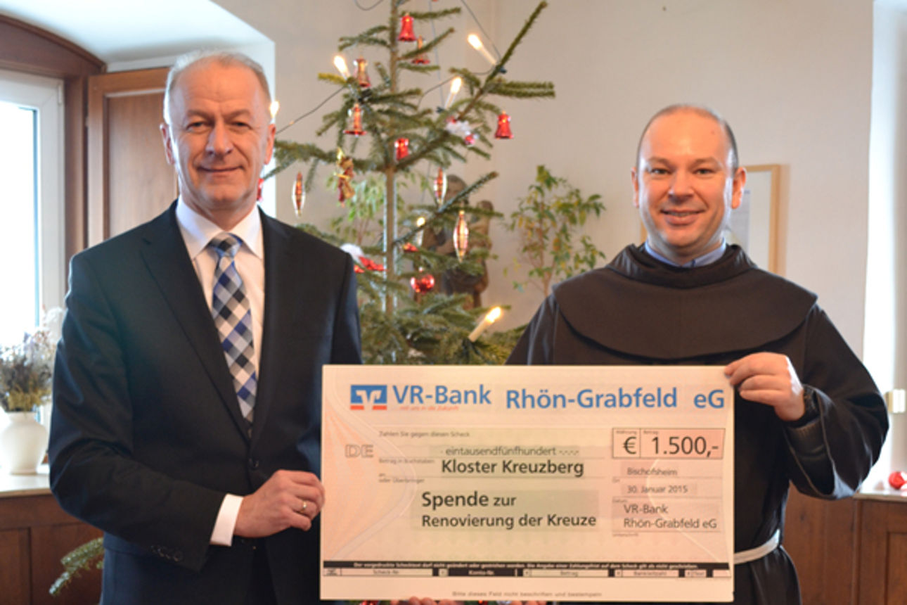 Vor einem Weihnachtsbaum stehen zwei Männer, die einen Spendenscheck in der Hand halten. Einer von ihnen trägt eine Kutte 