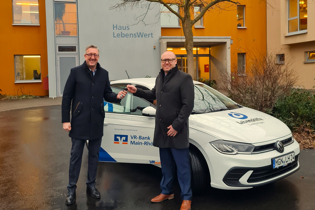 Zwei Männer stehen vor einem Gebäude mit der Aufschrift „Haus LebensWert“ und übergeben sich lächelnd einen Autoschlüssel