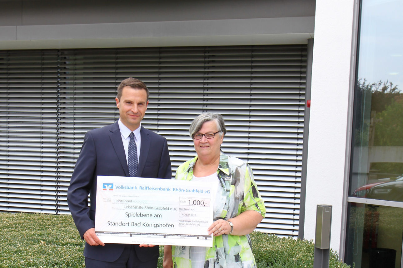 Ein Mann und eine Frau stehen vor einem Gebäude und halten einen Spendenscheck in der Hand. 