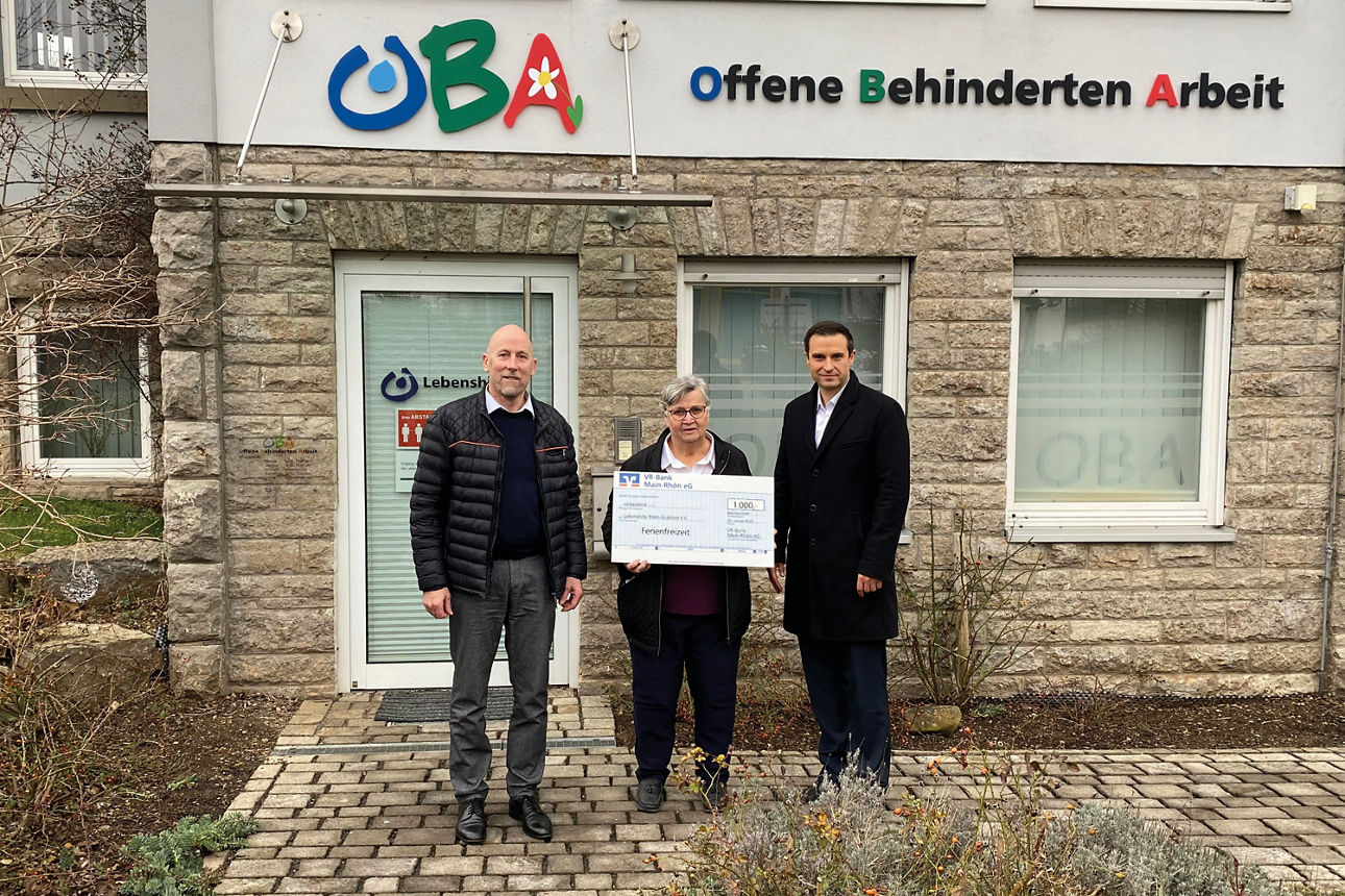 Drei Personen stehen vor einem Gebäude mit der Aufschrift „Offene Behinderten Arbeit“ und eine Frau hält einen Spendenscheck