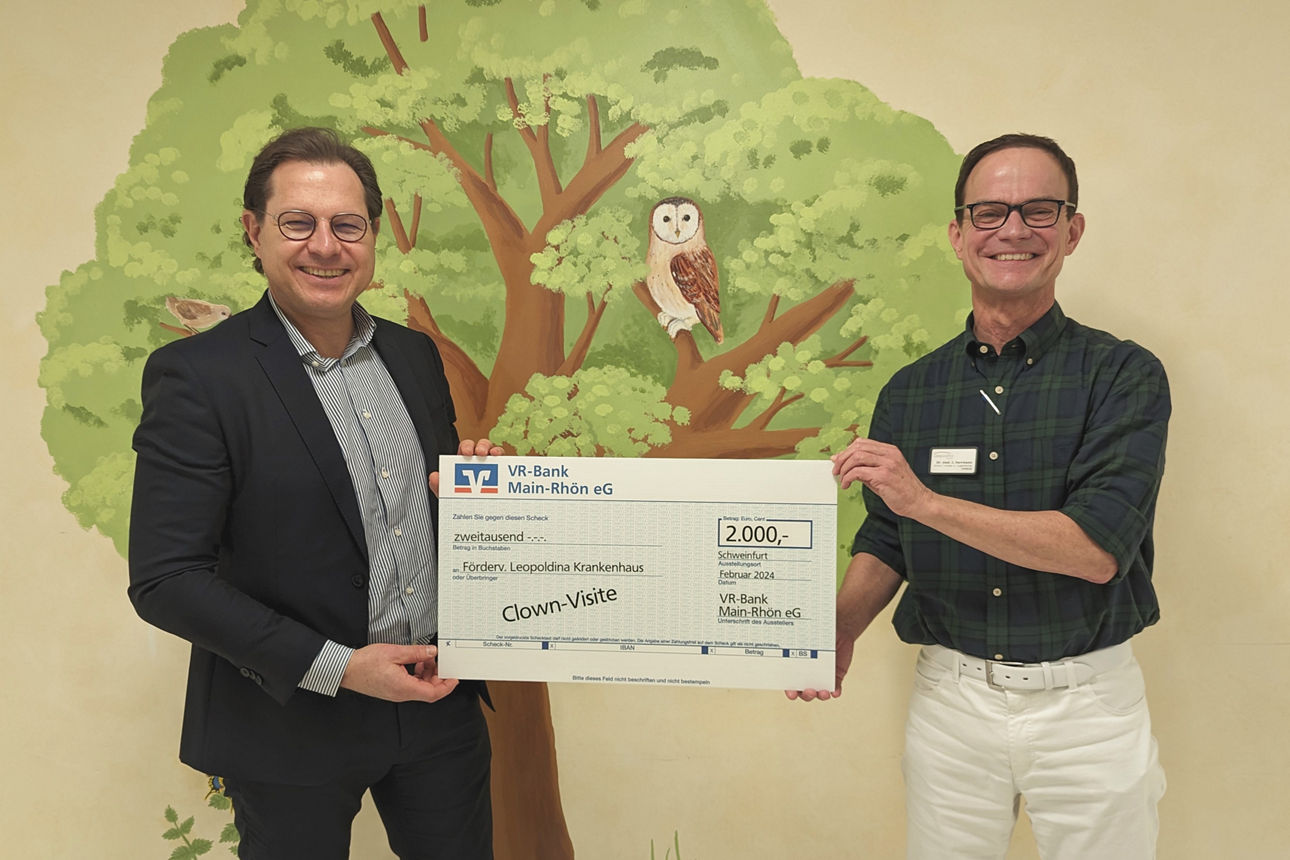 Zwei Männer stehen vor einer Wand mit einem Baumgemälde und halten gemeinsam einen großen symbolischen Scheck