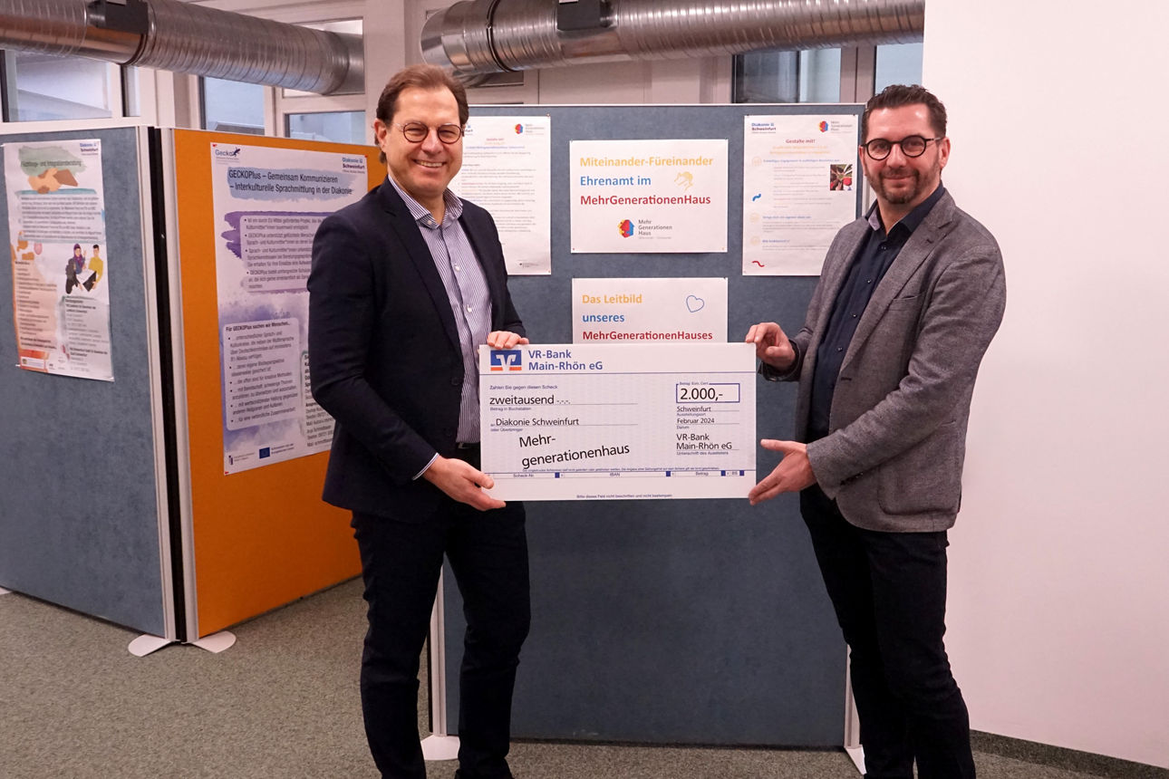 Zwei Männer stehen in einem Raum mit Infotafeln und halten gemeinsam einen großen symbolischen Scheck