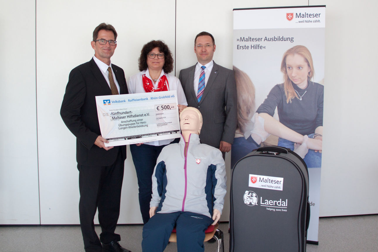 Zwei Männer und eine Frau stehen vor einer weißen Wand. Ein Mann und eine Frau halten einen Spendenscheck in der Hand