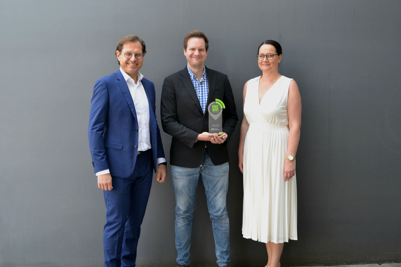 Auf dem Bild stehen drei Personen nebeneinander vor einer grauen Wand, der Mann in der Mitte hält eine Trophäe mit der Aufsch