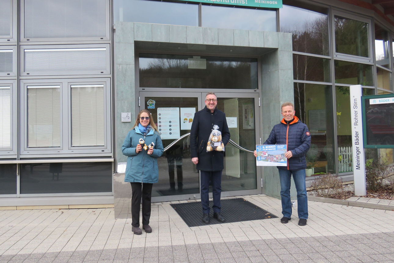 Vor dem Eingang des Gebäudes steht rechts ein Mann in der Mitte ein Mann mit Maskottchen und links eine Frau mit Kuscheltier