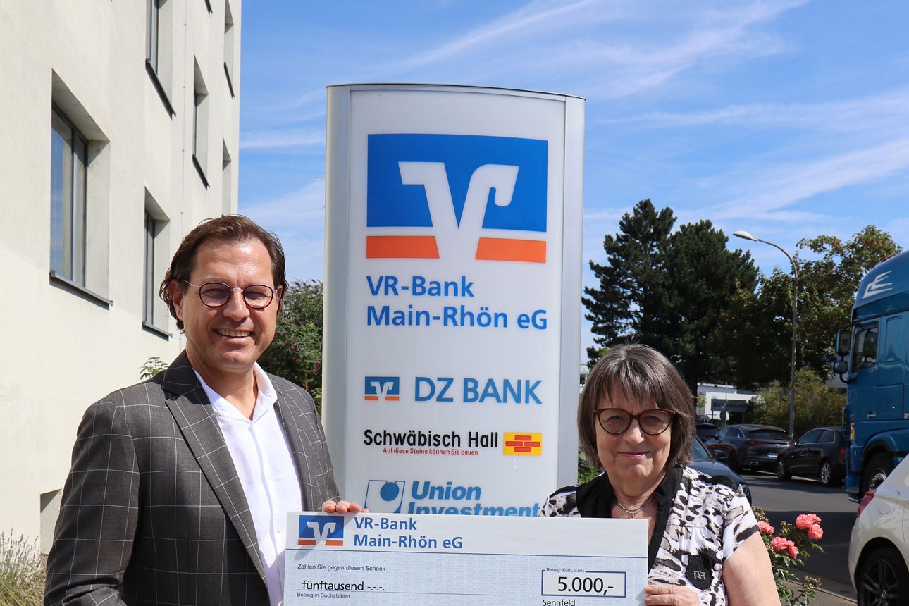 Auf dem Bild stehen ein Mann und eine Frau vor einem Schild der VR-Bank Main-Rhön, beide halten gemeinsam einen großen symbol