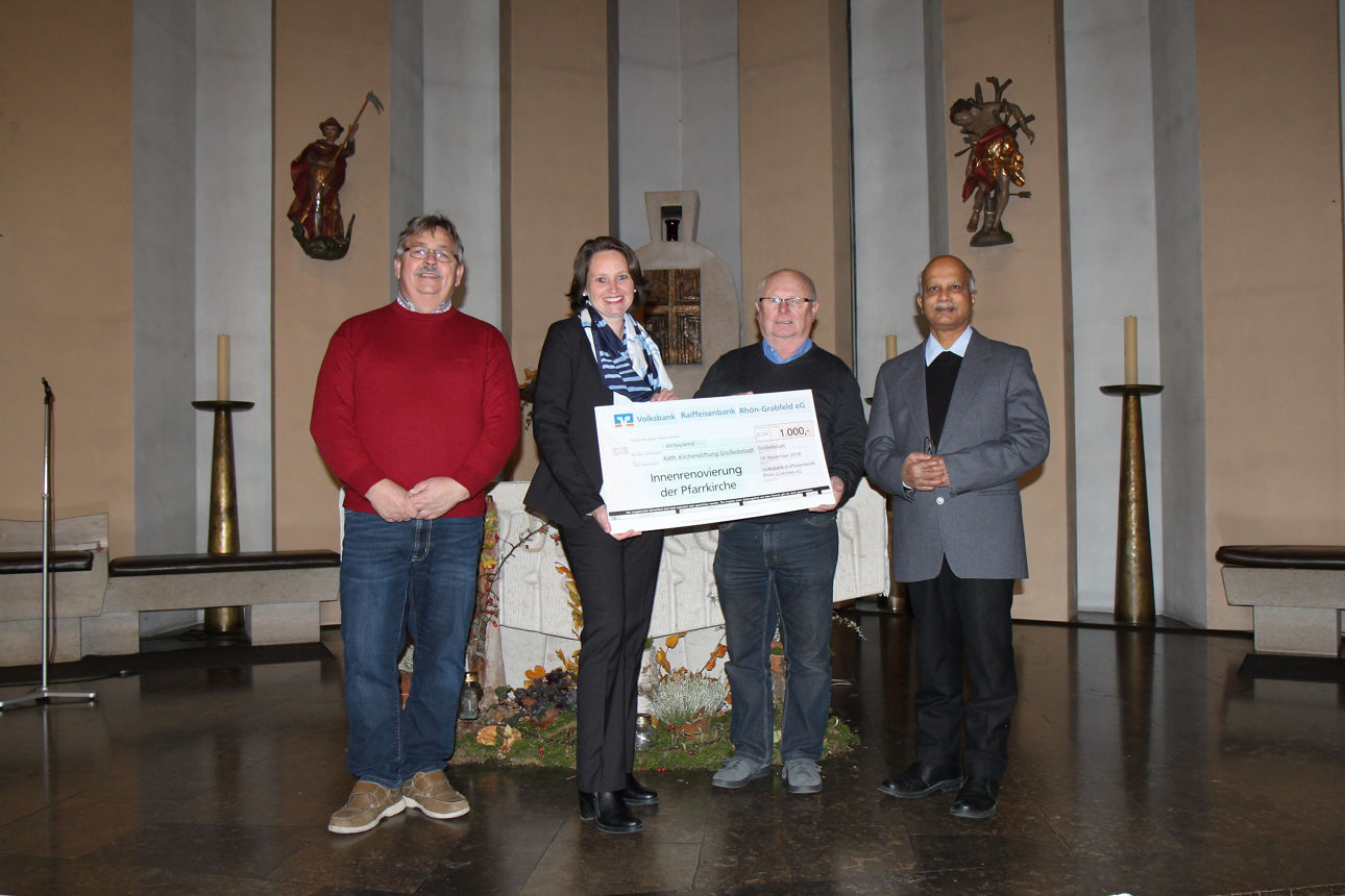 Drei Männer und eine Frau stehen vor einem Altar, die Frau und ein Mann hält einen Spendenscheck in der Hand