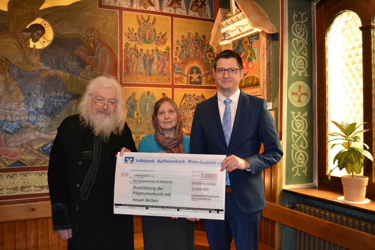 Zwei Männer und eine Frau stehen vor einer Wand und halten einen Spendenscheck 