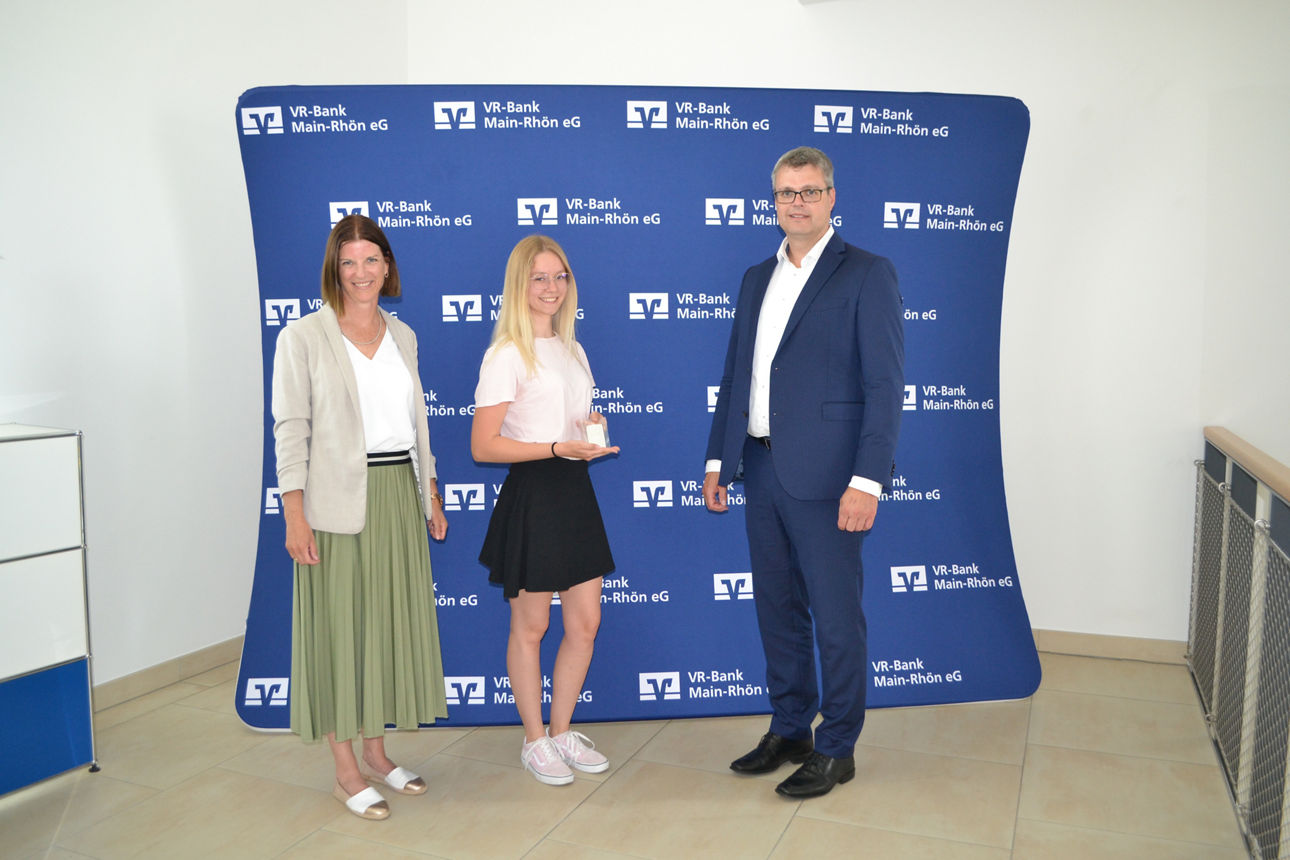 Drei Personen stehen vor einer Wand mit Logo der VR-Bank Main-Rhön, wobei die Frau in der Mitte einen Preis in der Hand hält