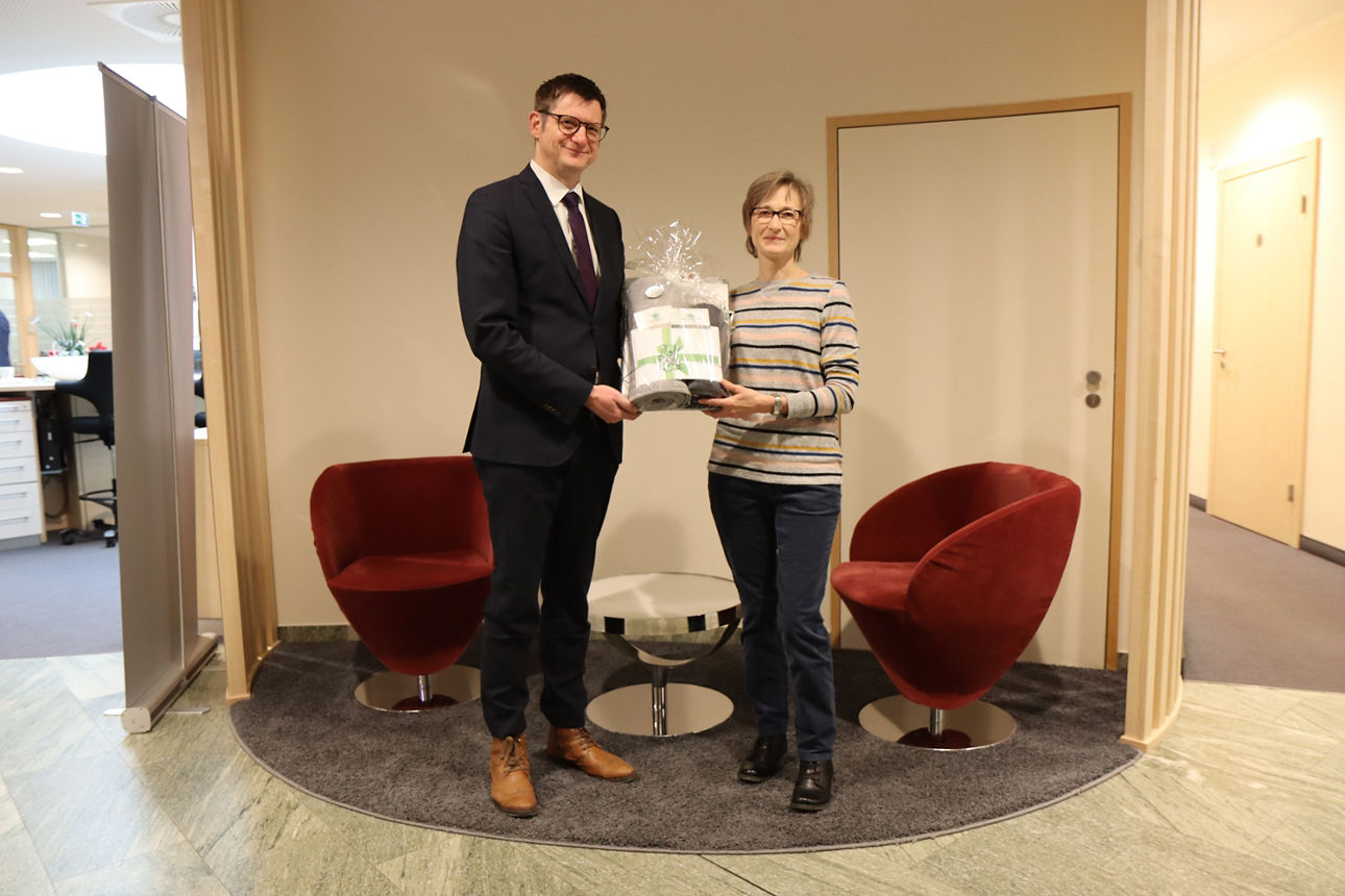 Vor einer weißen Wand vor der zwei rote Stühle stehen, übergibt ein Mann einen Präsentkorb an eine Frau 