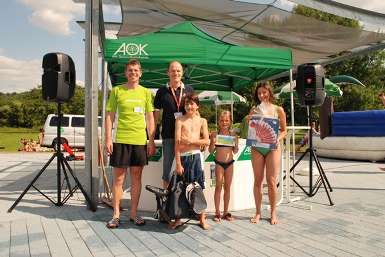 Fünf Personen, darunter zwei Erwachsene und drei Kinder in Badebekleidung, stehen vor einem grünen AOK-Zelt im Freien