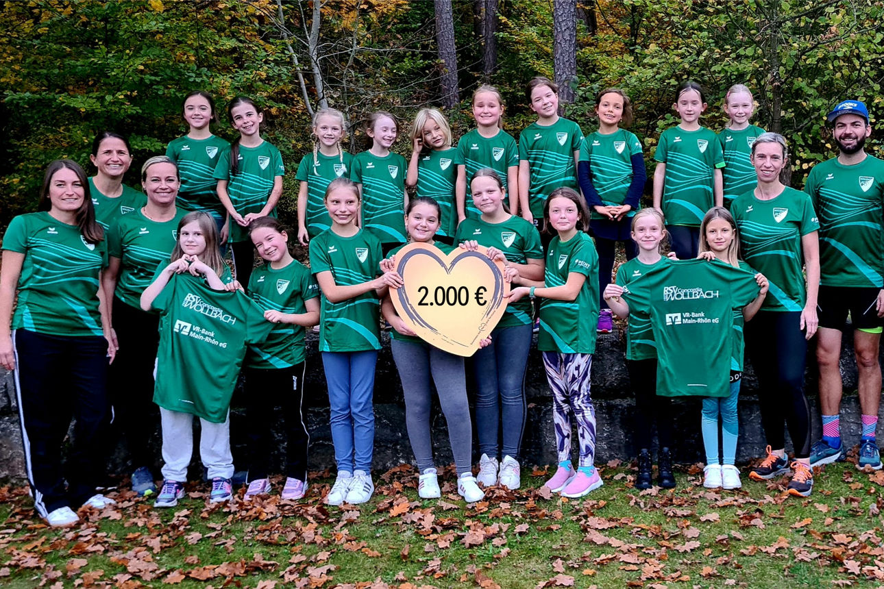 Eine Gruppe von Mädchen und Erwachsenen steht im Freien vor einer Waldkulisse; in der Mitte halten zwei Mädchen ein Herz 