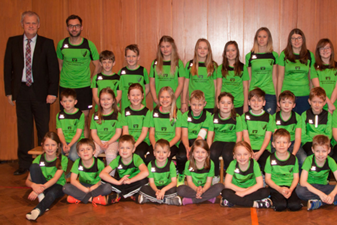 Vor einer Holzwand stehen und sitzen viele Kinder, die grüne Trikots tragen, ein Mann in Anzug steht links 