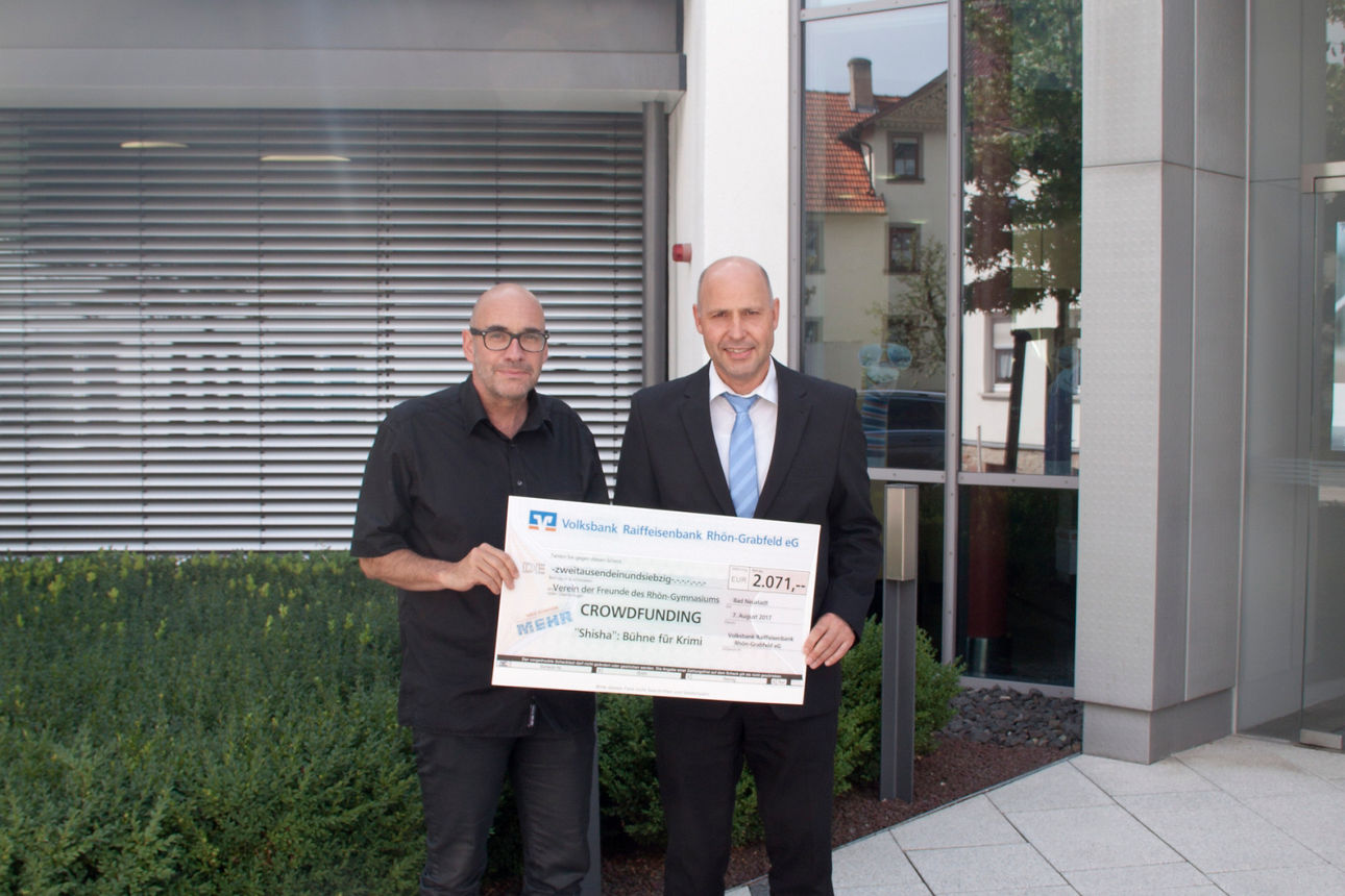 Zwei Männer stehen vor einem Gebüde und halten einen Spendenscheck in der Hand