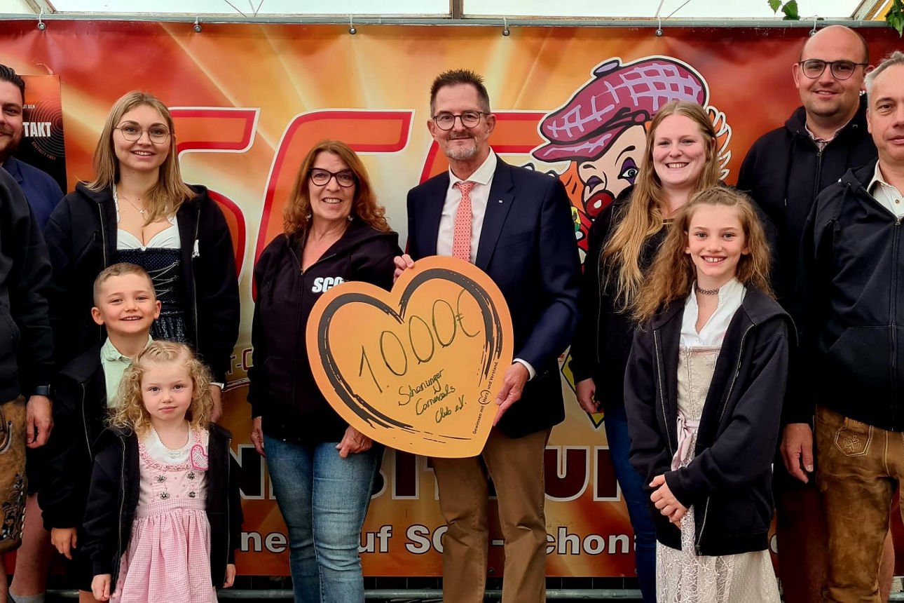 Eine Gruppe von Erwachsenen und Kindern steht zusammen vor einem farbenfrohen Hintergrund; eine Frau hält ein Schild 