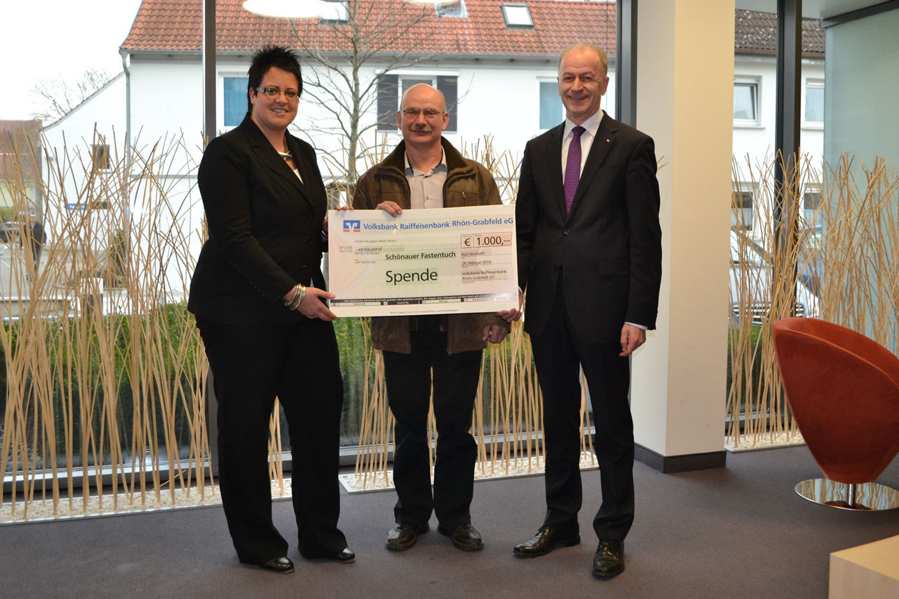 Zwei Männer und eine Frau stehen vor einem Fenster. Die Frau und ein Mann halten einen Spendenscheck