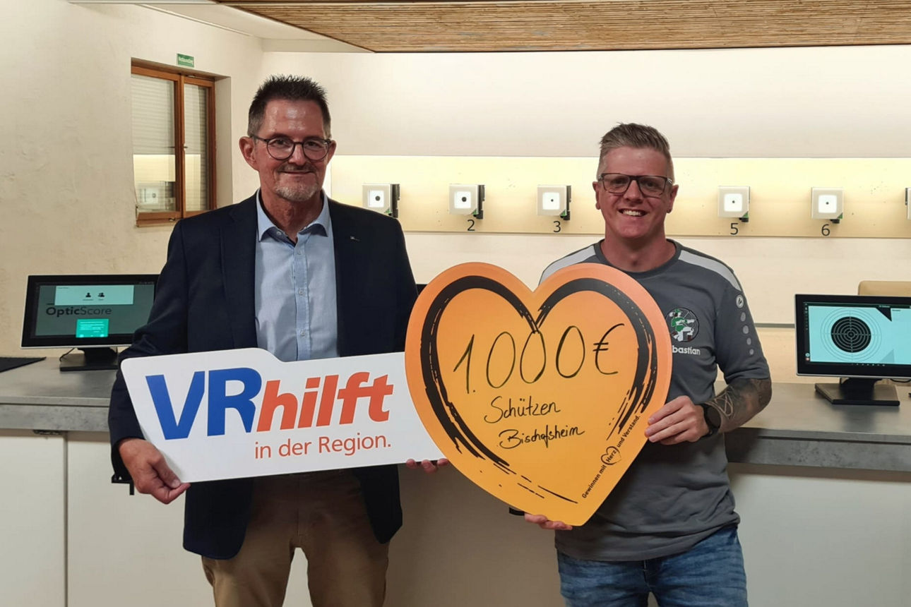 Zwei Männer stehen in einem Raum mit Schießständen; einer hält ein Schild mit der Aufschrift "VRhilft in der Region,"