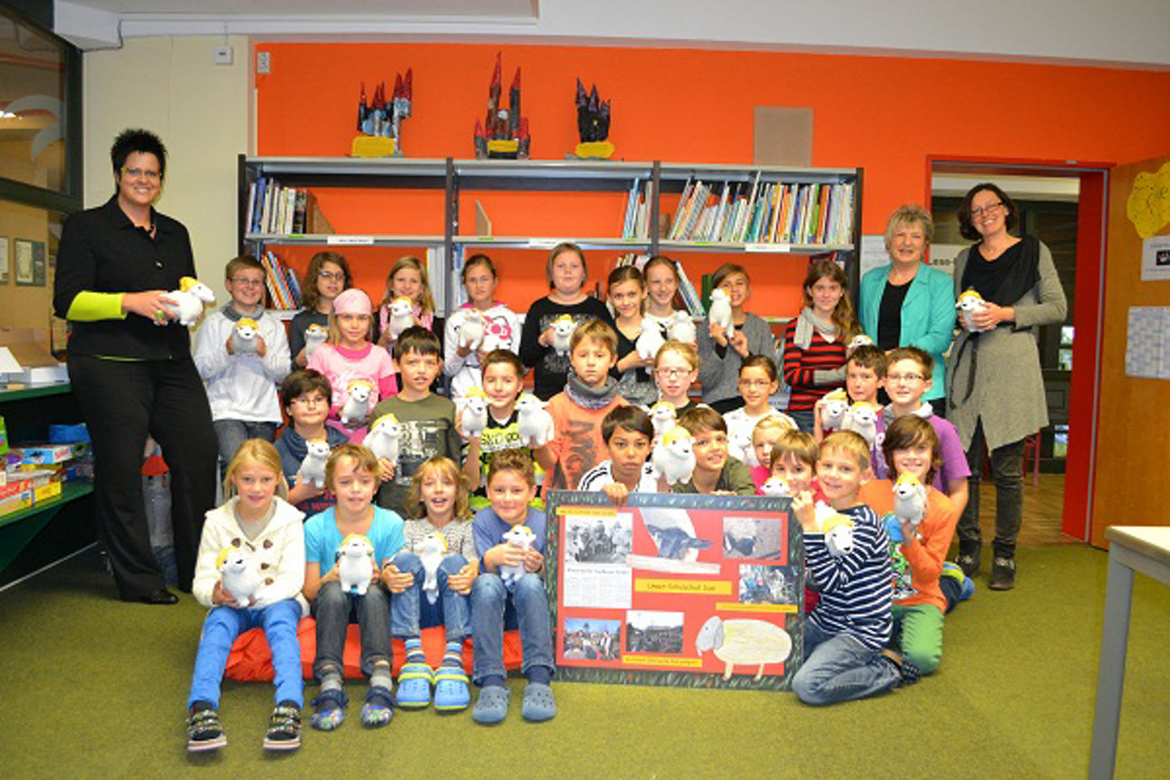 Das Bild zeigt eine Gruppe von etwa 30 Kindern die in einem bunt dekorierten Klassenraum stehen viele halten Stoffschafe