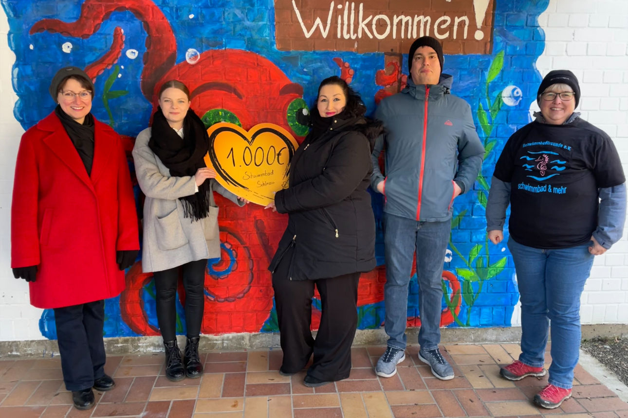 Fünf Personen stehen vor einer bunten Wand mit einem gemalten Oktopus und der Aufschrift „Willkommen!“; sie halten ein Herz