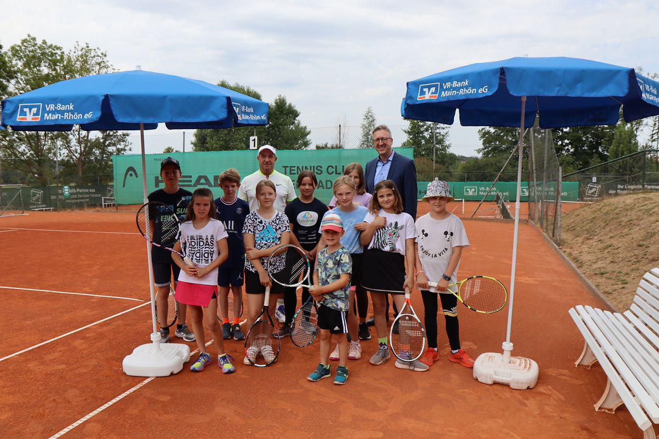 Auf dem Bild steht eine Gruppe von Kindern mit Tennisschlägern auf einem Sandplatz, begleitet von zwei Erwachsenen, unter zwe