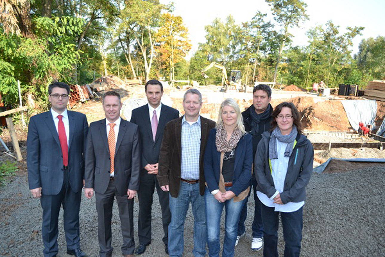 Sieben Personen stehen auf einer Baustelle mit Baumaterialien und Baggern im Hintergrund.