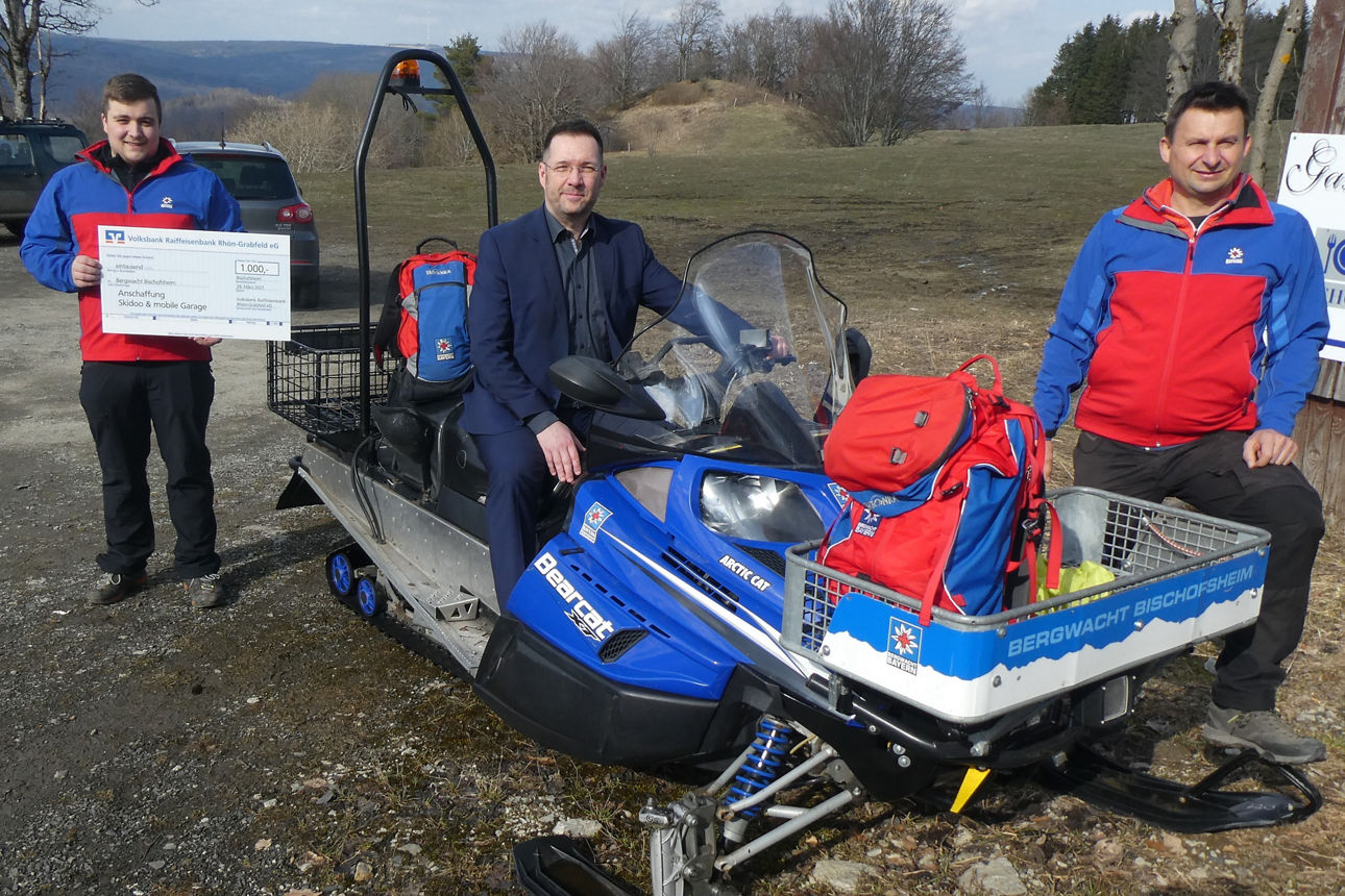 Ein Mann in der Mitte sitzt auf einem Schneemobil, rechts ein weiterer Mann, ein dritter steht links und hält einen Scheck