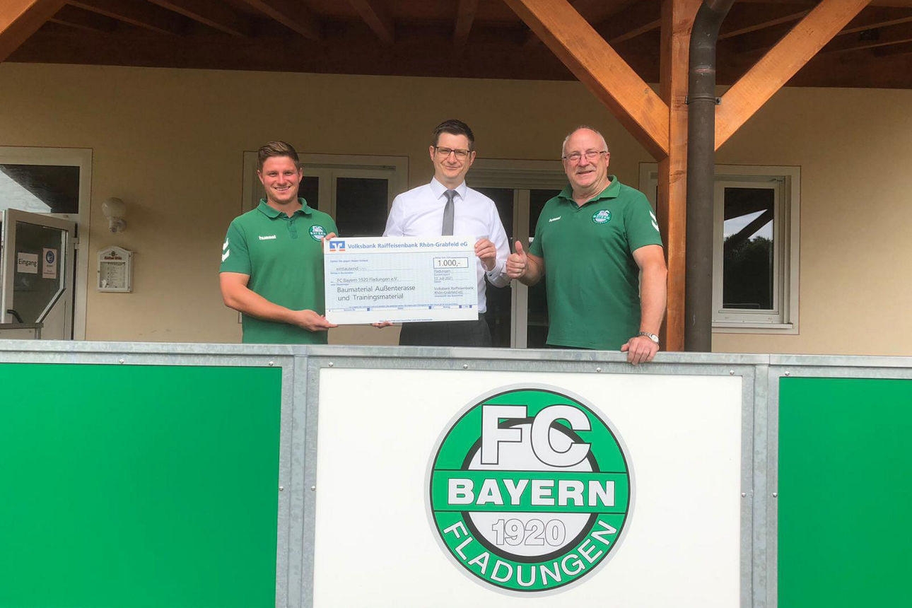 Drei Männer stehen hinter einer Bande und halten einen Gutschein in der Hand 