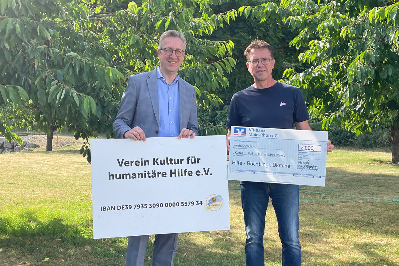 Zwei Männer halten auf einer Wiese ein Schild und einen Spendenscheck über 2.000 Euro für Ukraine-Flüchtlinge.