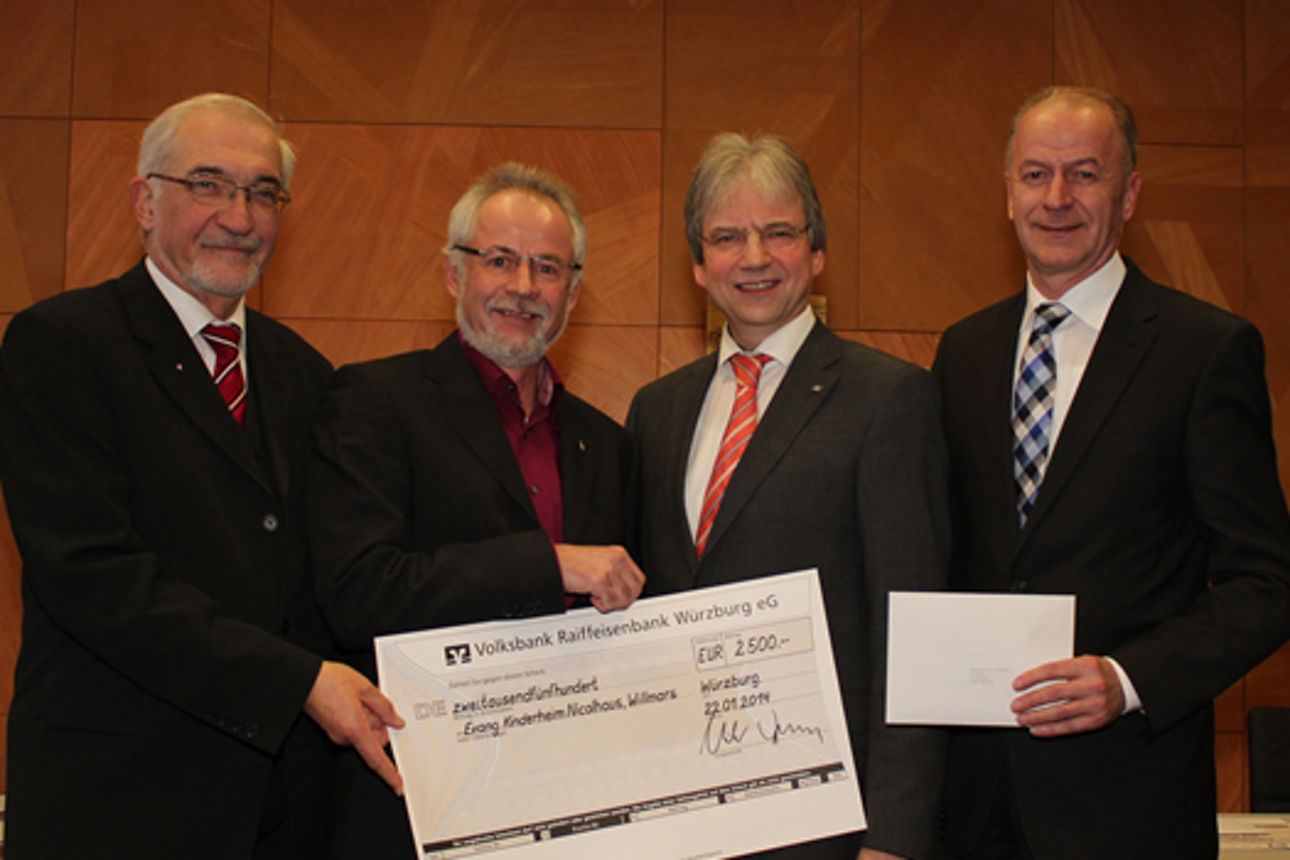 Vier Männer in Anzügen halten einen großen symbolischen Scheck über 2.500 Euro vor einer hölzernen Wand.