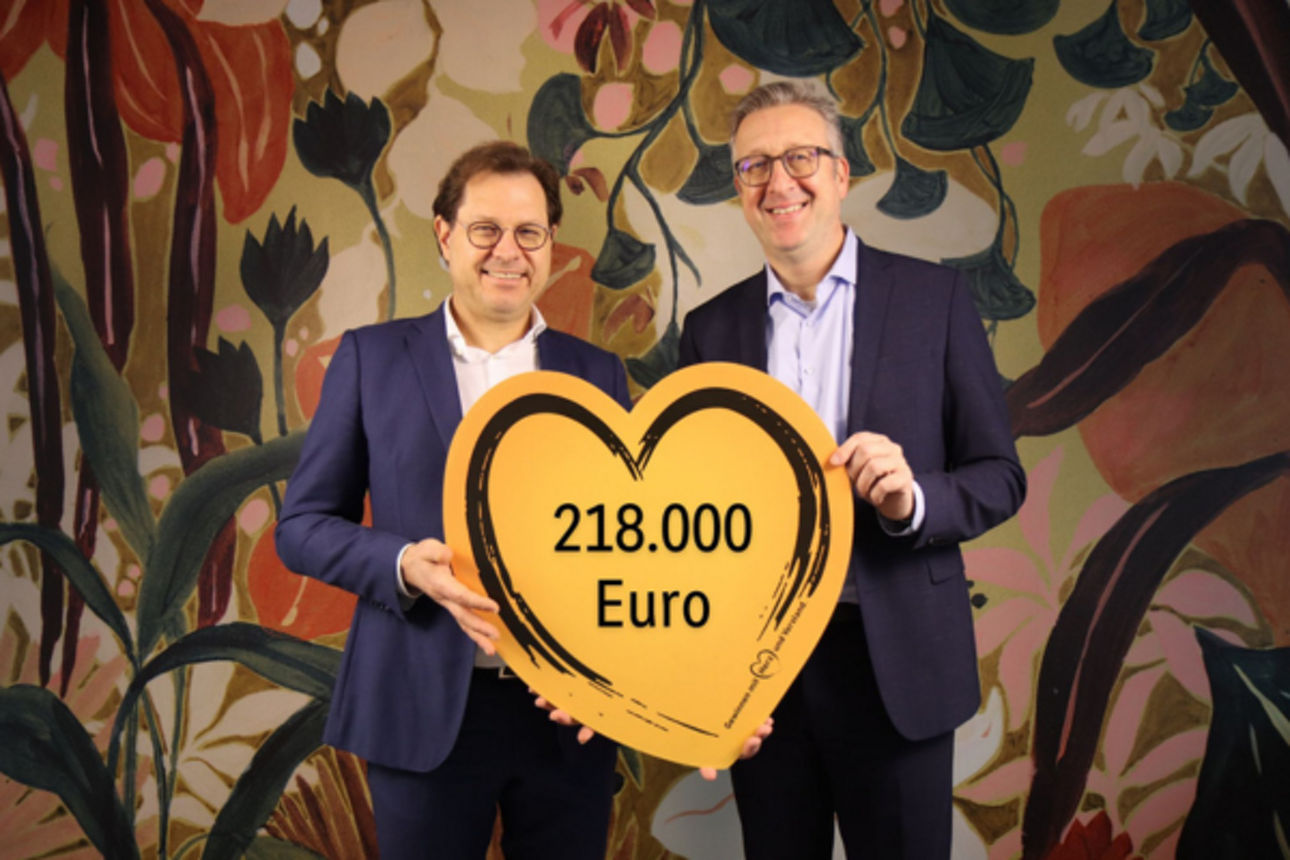 Vor einer Wand auf der Blumen aufgemalt sind stehen zwei Männer in Anzug, die ein gelbes Herz halten 