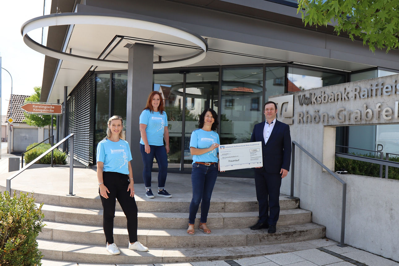 Drei Frauen und ein Mann stehen vor einem Bankgebäude. Der Mann und eine Frau halten einen Spendenscheck