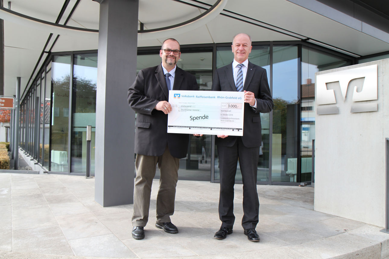 Zwei Männer stehen vor einem Bankgebäude und halten einen Spendenscheck in der Hand 